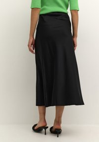 Schwarzer satinierter knöchellanger Rock mit glatter Textur und ausgestelltem Schnitt, kombiniert mit schwarzen Sandalen mit Absatz. Dunkler Hintergrund betont das Kleidungsstück.