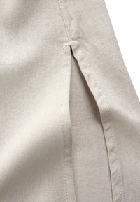 Primo piano di tessuto beige che mostra una cucitura laterale con una piccola apertura a spacco e dettagli di cucitura visibili.