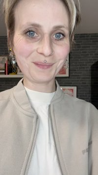 Tinkama bežinė bomber tipo striukė su užtrauktuku virš baltos marškinėlių. Sidabro spalvos auskarai žiede ir nosies žiedas, trumpi šviesūs plaukai, mėlynos akys.