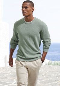 Hellgrüner, strukturierten Pullover mit Rundhalsausschnitt, kombiniert mit beigen Hosen. Der Pullover hat gerippte Bündchen und Saum und besteht aus einem weichen Strickmaterial.