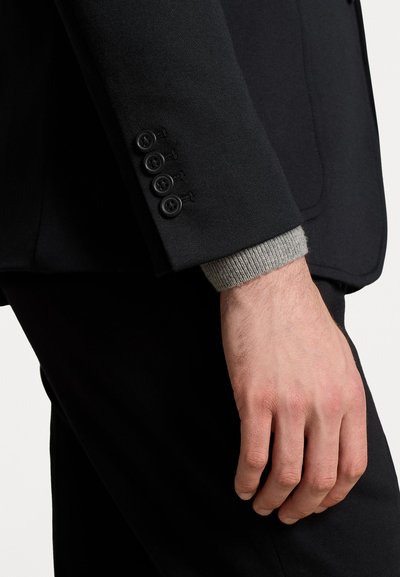 Blazer noir avec une coupe ajustée, doté de quatre boutons noirs sur le poignet. La manche comprend un poignet en tricot gris en dessous.