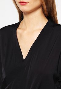 Blouse noire à enfiler avec un col en V et une texture douce et lisse. Le design est simple et épuré, sans motifs visibles ni accessoires.