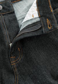 Jeans din denim închis, desfăcute, care arată fermoarul metalic, cusătura portocalie, materialul interior alb și zona butonului cu o deschidere vizibilă pentru buton.