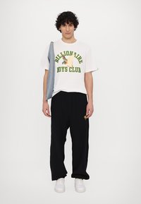 Λευκό βαμβακερό T-shirt με πράσινο κείμενο "Billionaire Boys Club", σε συνδυασμό με μαύρα παντελόνια φόρμας και λευκά αθλητικά παπούτσια. Τζιν μπουφάν κρεμασμένο στον ώμο.