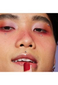 Närbild av ett ansikte med mörkt hår, iförd rosa ögonskugga med röda accenter, dekorativa ansiktspiercingar och glänsande nakna läppar. Röda nagellack.