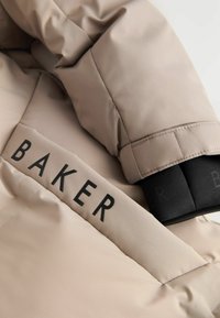 Beige gepolsterte Jacke mit schwarzem "BAKER"-Logo am Saum. Verfügt über eine glatte Textur und strukturierte Ärmel mit weichen, elastischen Bündchen.