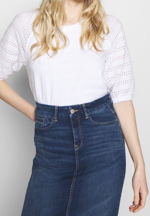 Frau mit langem blondem Haar, die ein weißes Top mit Lochstickmuster und einen hoch taillierten dunkelblauen Denimrock trägt, eine Hand in der Hüfte.