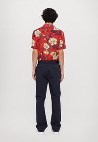 Camisa corta de flores rojas con acentos en color crema y azul, combinada con pantalones oscuros navy. La camisa tiene un corte relajado y un cuello estándar.