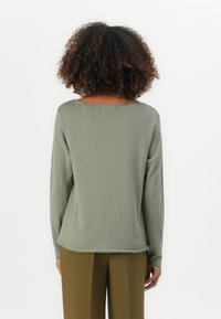 Camicia a maniche lunghe color verde oliva con collo rotondo, texture morbida e orlo curvo, abbinata a pantaloni khaki.