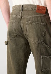 Olive green cargo pants with a tailored fit, featuring vertical seams, two back pockets, and a leather logo patch on the waistband.