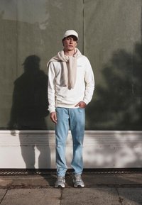 Felpa bianca, jeans azzurri chiari, maglione beige annodato attorno al collo, sneakers grigie e berretto beige, in piedi su uno sfondo verde.