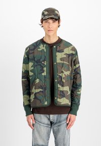 Νέος άνδρας που φοράει πράσινο και καφέ καμουφλάζ μπουφάν με επένδυση και ταιριαστό καπέλο, σκούρο πουκάμισο και ανοιχτόχρωμα μπλε τζιν, στέκεται μπροστά σε λευκό φόντο.