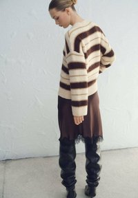 Suéter de mezcla de lana a rayas en crema y marrón, combinado con un vestido de satén marrón con ribete de encaje y botas ajustadas de cuero negro.