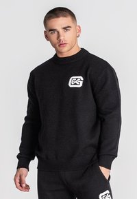 Junger Mann mit kurzem Haar, der einen schwarzen Sweatshirt und passende Hosen trägt, beide mit einem weißen Logo auf der Brust und dem Oberschenkel.