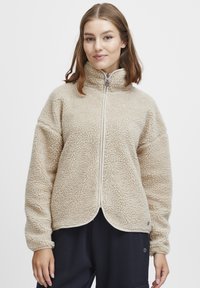 Beige fleece zip-up jakke med høy krage, teksturert overflate, sidelommer og kontrasterende glidelåsdetaljer. Glatt, avrundet kant.