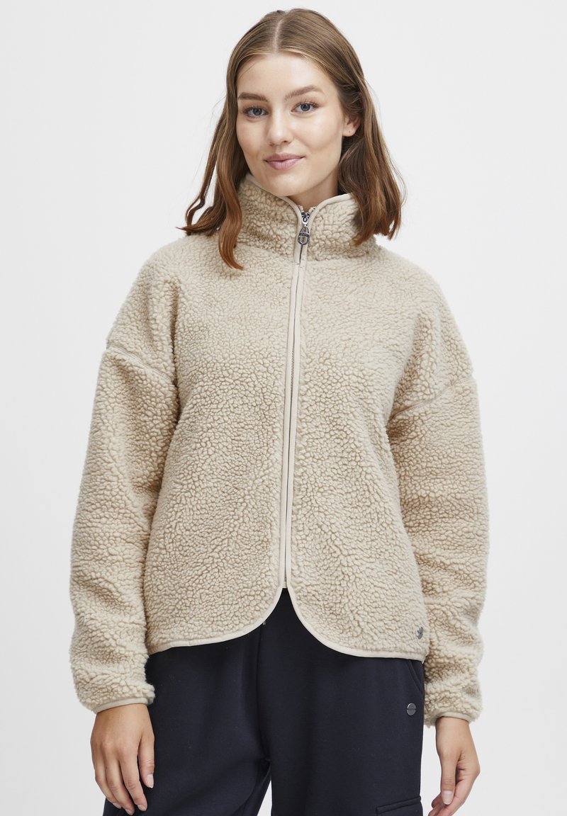 Beige fleece zip-up jakke med høy krage, teksturert overflate, sidelommer og kontrasterende glidelåsdetaljer. Glatt, avrundet kant.