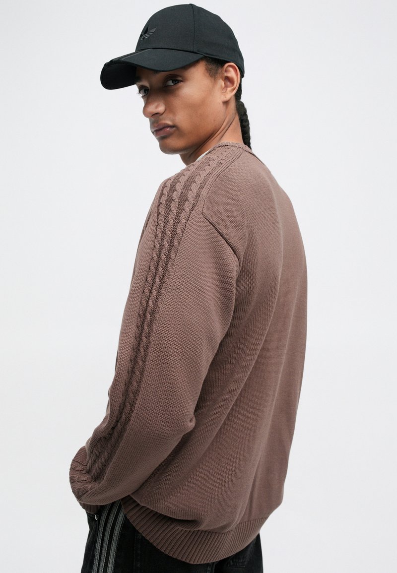 Brown knit sweater with ribbed cuffs and textured side details. Worn with black pants and a black cap. Smooth fabric and relaxed fit.