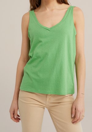 Kobieta ubrana w zielony top bez rękawów z dekoltem w serek oraz beżowe spodnie, stojąca na jasnym, jednolitym tle, ukazująca tułów i ramiona.