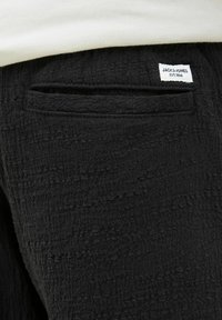 Crne teksturirane hlače s vidljivim stražnjim džepom i bijelom etiketom brenda na kojoj piše "JACK & JONES EST. 1990." Mekana tkanina.