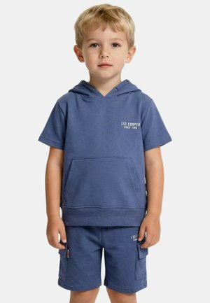 Jeune garçon aux cheveux blonds courts portant un haut à capuche à manches courtes bleu et un short assorti avec des poches avant, debout devant un fond uni.