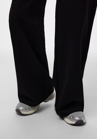 Črne široke hlače s širokimi hlačnicami delno pokrivajo srebrne superge z mrežastimi in gumijastimi poudarki; teksturirana tkanina z gladkim zaključkom.