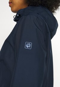 Veste imperméable bleu marine avec capuche ajustée, décorée d'un logo carré avec une empreinte de patte sur la manche gauche. Texture lisse, design minimaliste.