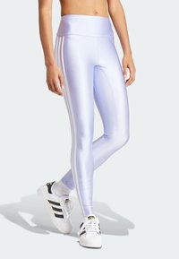 Person som bär glansiga lavendelfärgade tights med vita sidostripes och vita Adidas sneakers med svarta ränder, stående med korsade ben.