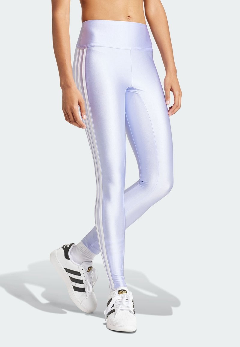 Person som bär glansiga lavendelfärgade tights med vita sidostripes och vita Adidas sneakers med svarta ränder, stående med korsade ben.