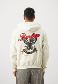 Sudadera crema con un gran gráfico de un águila y el texto "Benfica" en rojo en la parte posterior. Incluye una pancarta con "E Pluribus Unum" debajo.
