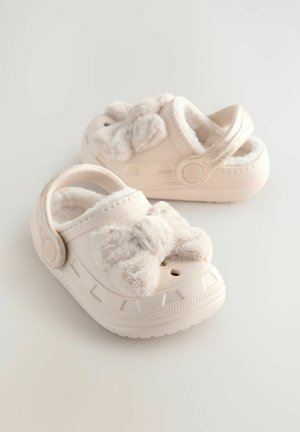 Beige Clogs mit weichem, flauschigem Schleifendetail an der Vorderseite. Aus leichtem Material gefertigt, mit einer strukturierten Sohle und verstellbarem Riemen hinten.