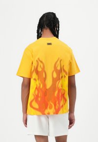 Tricou din bumbac galben, cu un design grafic de flacără în culori portocalie și roșie pe spate, având un mic logo Nike la guler.