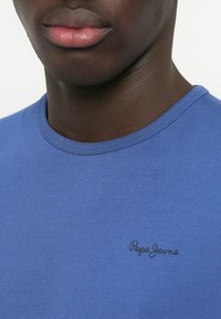 Modrá bavlnená tričko s okrúhlym výstrihom a krátkymi rukávmi. Obsahuje čierne logo "Pepe Jeans" na ľavej strane pri hrudníku.