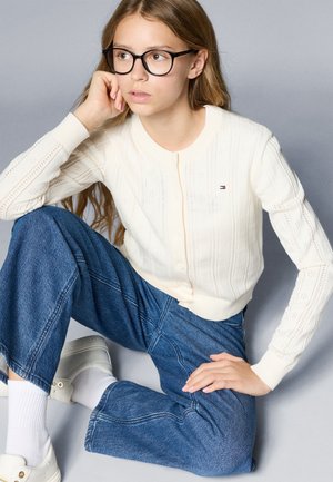 Vit kabelstickad cardigan med ribbade ärmslut och knäppning fram. Kombinerad med lösa denimjeans och vita sneakers. En enkel grå bakgrund.