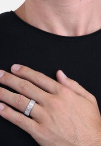 Srebrny pierścionek z grawerowanym wzorem, gładkim wykończeniem i płaskim kształtem obrączki. Dłoń spoczywa na czarnym tle z tkaniny.