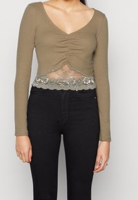 Kvinna iklädd en olivgrön långärmad topp med rynkad framdel och spetskant, kombinerad med höga svarta jeans.