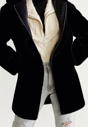 Veste noire duveteuse zippée portée ouverte sur un pull crème en maille torsadée et un jean gris clair déchiré.