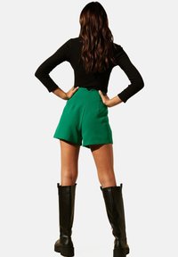 Shorts verts taille haute en tissu lisse, présentant une silhouette épurée. Associés à un haut noir à manches longues en côte et des bottes noires ajustées jusqu'aux genoux.