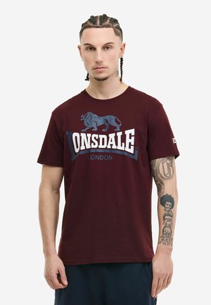 Man with braided hair wearing a burgundy Lonsdale London t-shirt and dark pants, showing tattoos on his right forearm.