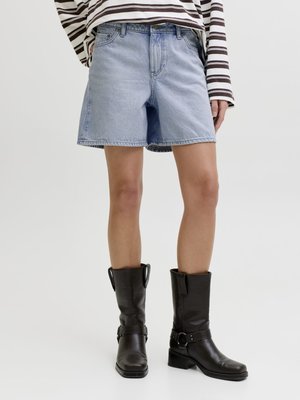 Osoba oblečená v svetlomodrých rifle krátkych nohaviciach, čiernych kožených čižmách po stred lýtok a v pruhovanom tričku s dlhým rukávom stojaca na jednoduchom pozadí.