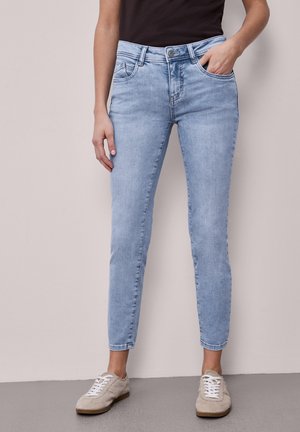 Hellblaue Slim-Fit-Jeans, getragen mit beigen Sneakern, eine Hand in der Tasche, die andere entspannt seitlich am Körper, vor einfachem Hintergrund.