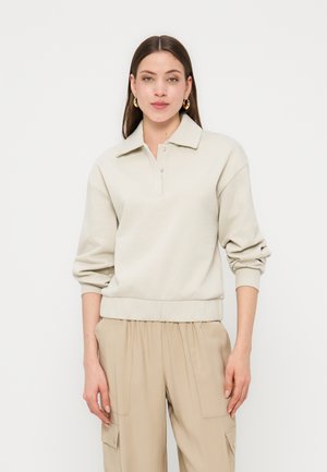 Hosszú sötét hajú nő arany karika fülbevalóval, bézs színű galléros pulóvert és laza bézs cargo nadrágot viselve, fehér háttér előtt áll.