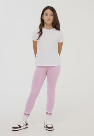 T-shirt bianco a maniche corte, leggings rosa chiaro e sneaker. I leggings aderiscono perfettamente; i calzini presentano strisce rosa, bianche e blu ai polsini.