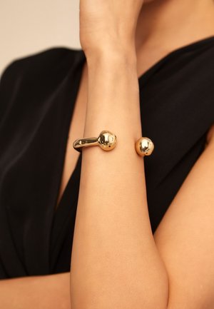 Bracelet manchette de couleur dorée avec une finition lisse et polie, orné de deux accents sphériques, porté sur un bras nu contre un fond en tissu noir.