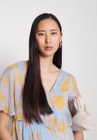 Vestido floral em azul pálido com padrões amarelos, design em decote em V e mangas em camadas. A segurar uma bolsa tecida em bege com textura.