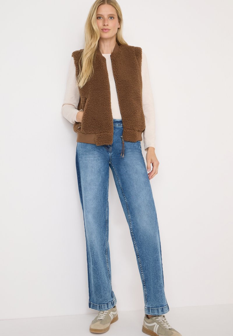 Brown faux shearling vest with a front zipper, side pockets, and ribbed hem, worn over a light sweater and paired with wide-leg blue jeans.