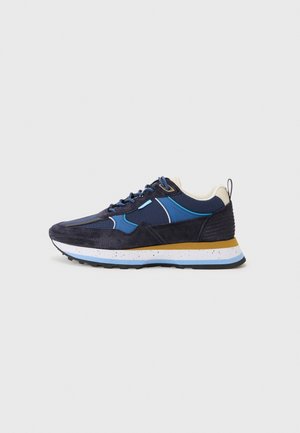 Donkerblauwe en zwarte sneaker met blauwe accenten, witte en gele gespikkelde zool en zwart profiel, getoond vanaf de zijkant op een witte achtergrond.