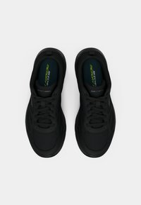 Ténis desportivos pretos com parte superior em rede, biqueira arredondada, fecho de atacadores e palmilhas de espuma com memória. Detalhe notável do logótipo na língua.