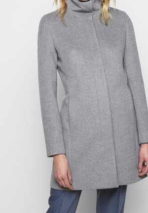 Personne portant un manteau en laine gris clair ajusté avec un col haut et des manches longues sur un pantalon bleu texturé.