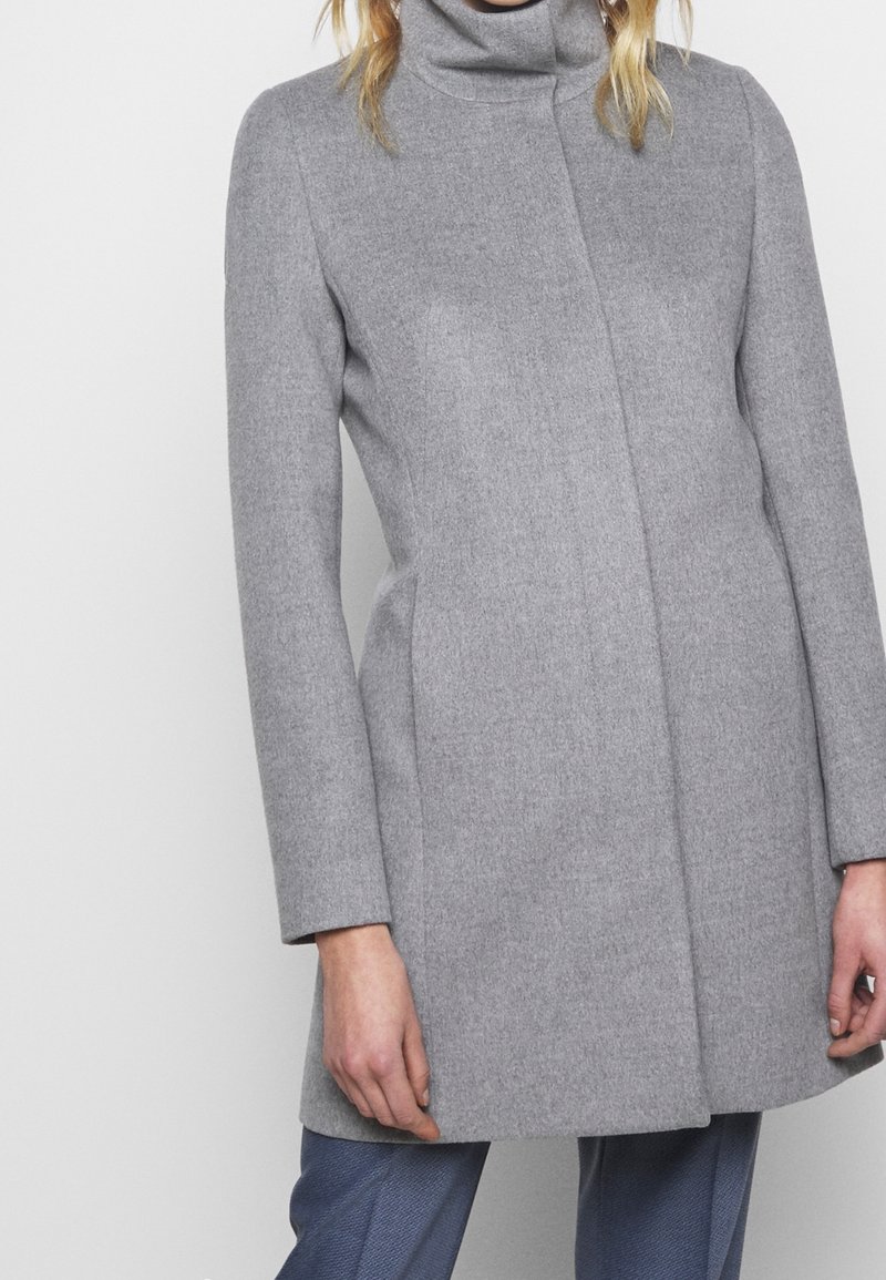 Personne portant un manteau en laine gris clair ajusté avec un col haut et des manches longues sur un pantalon bleu texturé.