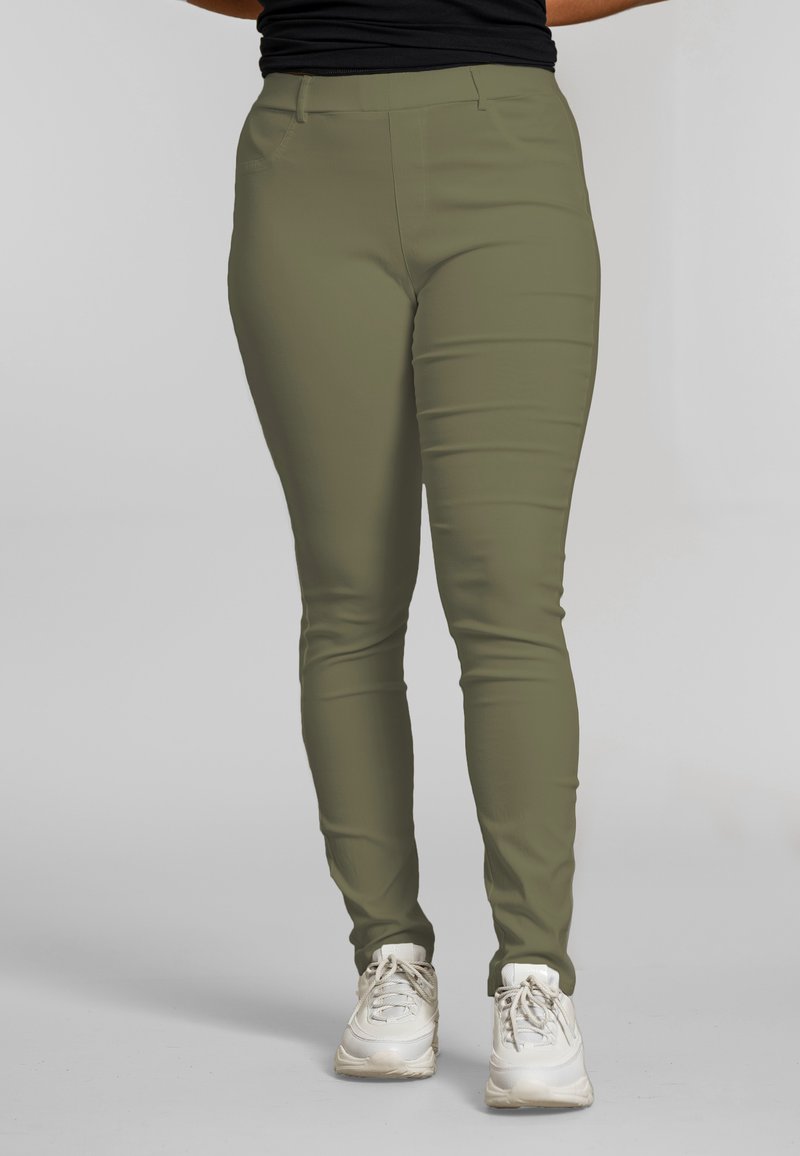 Frau, die olivgrüne, figurbetonte Hose und weiße Turnschuhe trägt, steht vor einem einfarbigen grauen Hintergrund.